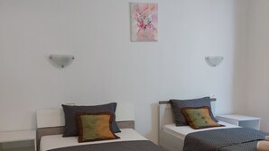 A hotel room with two beds, each with a headboard, a bedside table, and a wall-mounted light. There is a painting on the wall above the beds.