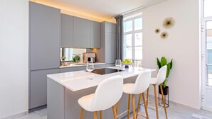 Une cuisine moderne dotée d’un îlot central, de tabourets de bar blancs et d’une grande fenêtre qui laisse entrer la lumière naturelle.