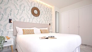 Un lit bien fait, avec du linge de lit blanc et des coussins à motifs, une table de chevet avec une plante et un miroir fixé au mur.