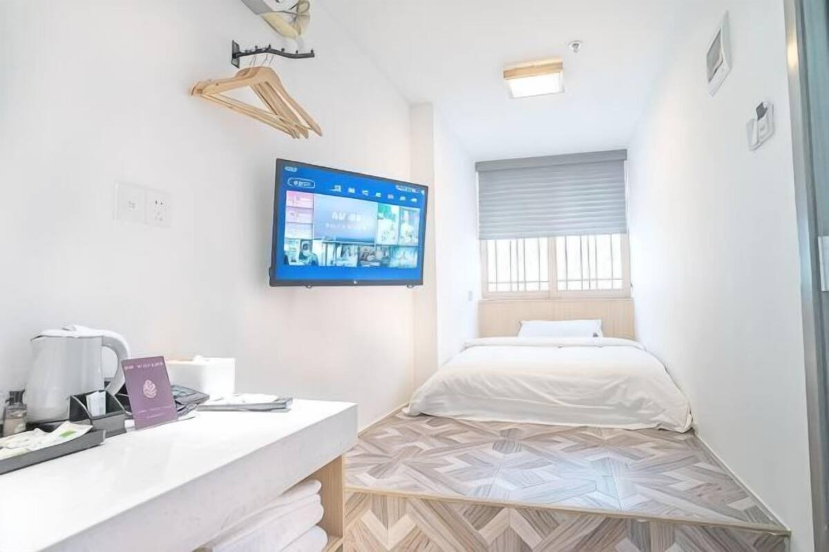 A hotel room with a bed, a desk with a laptop, a TV mounted on the wall, and a window with blinds.