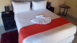 Un lit bien fait, avec du linge de lit blanc et un élément décoratif rouge, une tête de lit rembourrée de couleur grise, deux tables de chevet avec des lampes et une petite table sur laquelle se trouve un plateau avec divers objets.