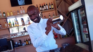 Un barman prépare un cocktail dans un bar où l’on peut voir, derrière lui, des étagères remplies de bouteilles d’alcool et un réfrigérateur.