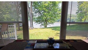 Una habitación con un ventanal grande, una mesita con un florero y vista a un árbol y césped afuera.