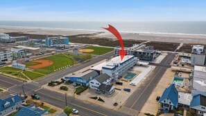 An aerial view of a coastal town with a prominent building marked by a red arrow, a baseball field, and a beach in the background.