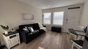 A living room with a black leather sofa, a white side table, a small bench, and a dining table with chairs. There are two windows with blinds, a door, and a wall-mounted sign. - Sunny Wildwood Crest condo! Perfect spot, 1 block to beach ☀️🌊 (Wildwood)