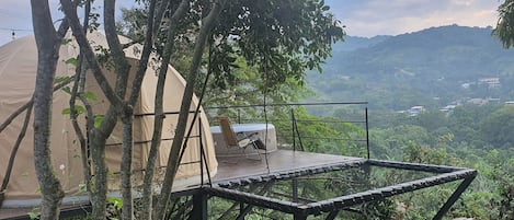 A tented accommodation with a platform and a chair, surrounded by trees and a view of a hilly landscape.