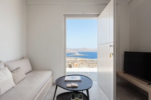 A modern living room with a sofa, a small round table with a book and two stones, a TV on a stand, and a view of the sea and mountains through an open door.