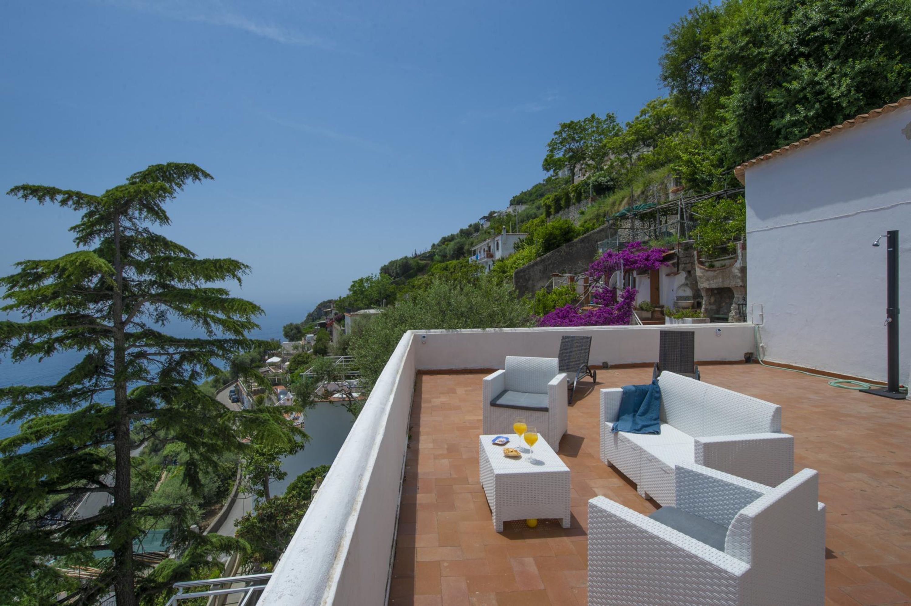 A rooftop terrace with white wicker furniture, a small table with a plate and glass, and a view of the sea and greenery.