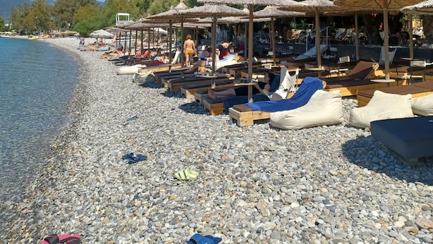 Una playa con piedras, tumbonas y sombrillas, un cielo azul despejado y árboles al fondo.
