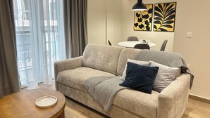 Un salon comprenant un canapé, une table ronde en bois, une table à manger avec des chaises et des œuvres d’art accrochées au mur.