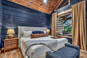 A bedroom with a bed, bedside tables, a lamp, a window with curtains, and a view of greenery outside.