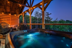 A hot tub under a wooden roof with string lights, surrounded by chairs and a view of trees.