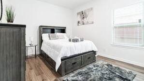 Ein Schlafzimmer mit Bett, Nachttisch, Lampe und einem Fenster mit Jalousien.