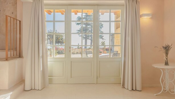 A room with a large window with white curtains, a wooden ceiling, and a small table with a vase.