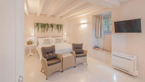A bedroom with a bed, two wicker chairs, a bedside table, a TV, and a wall-mounted heater.