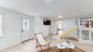 A spacious living area with a wooden floor, a white door with a window, a television mounted on the wall, and a yellow slide. - The Playmakers Funhouse: HOF Village (Canton)