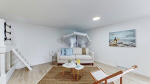 A modern living room with a white sofa, a small round table, a woven rug, a wall-mounted umbrella, and a painting of a beach scene. - The Playmakers Funhouse: HOF Village (Canton)
