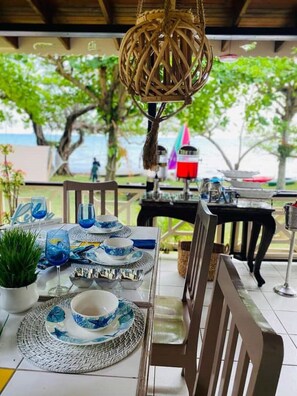 A table set for dining with blue and white tableware, a hanging wicker light fixture, and a view of the sea and trees in the background.