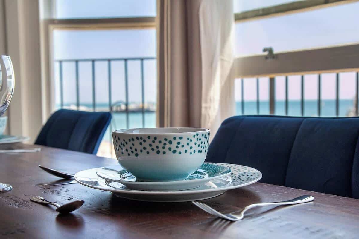 A dining table with a bowl and plate set, a spoon, and a fork. The table is in a room with a view of the sea through a window.