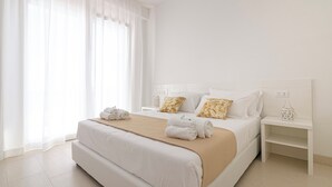 A modern hotel room with a large bed, white bedding, and a beige throw blanket. There are two bedside tables with decorative items and a window with sheer curtains. - Rivazzurra Homes - 24 (Litorale Nord)