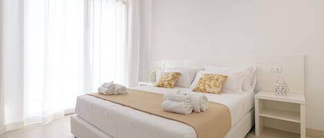 Quarto de hotel moderno com uma cama grande, roupa de cama branca e um cobertor bege. Há duas mesas de cabeceira com objetos decorativos e uma janela com cortinas leves.