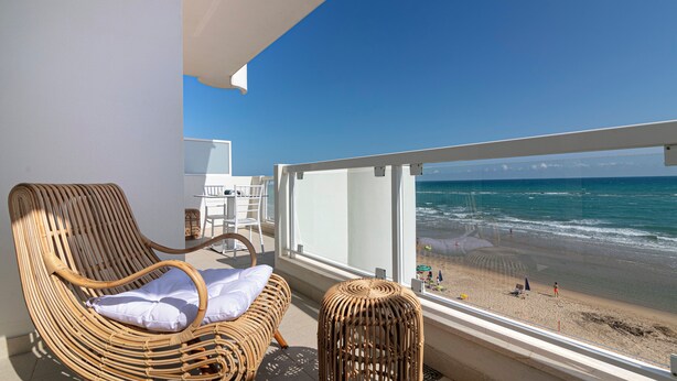 A balcony with wicker chairs and a small table, overlooking a beach with people and a clear blue sky. - Rivazzurra Homes - 24 (Litorale Nord)
