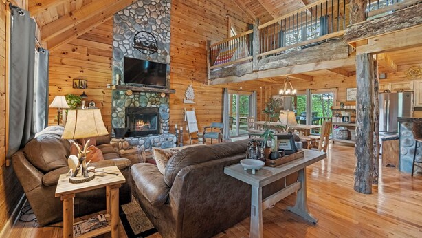A spacious log cabin interior with a stone fireplace, leather furniture, and a balcony overlooking the outdoors.