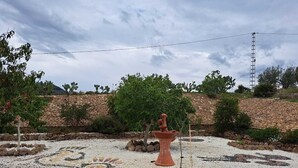 A garden with a central terracotta fountain, surrounded by pebbles and rocks, with cacti and trees in the background. - Casa de Campo. Confortable y Comoda (Alozaina)