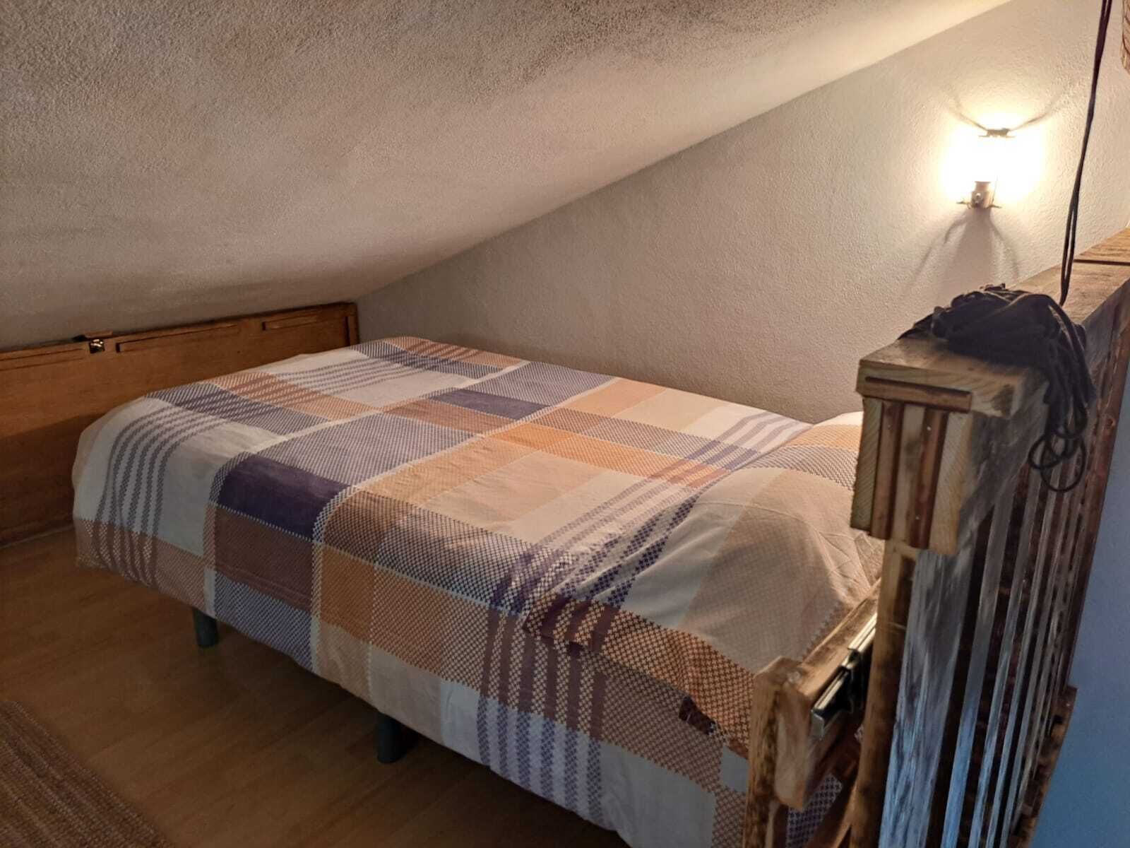Une petite chambre mansardée, chaleureuse et accueillante, avec un plafond en pente, une tête de lit en bois, une couvre-lit matelassé et une seule lampe de chevet.