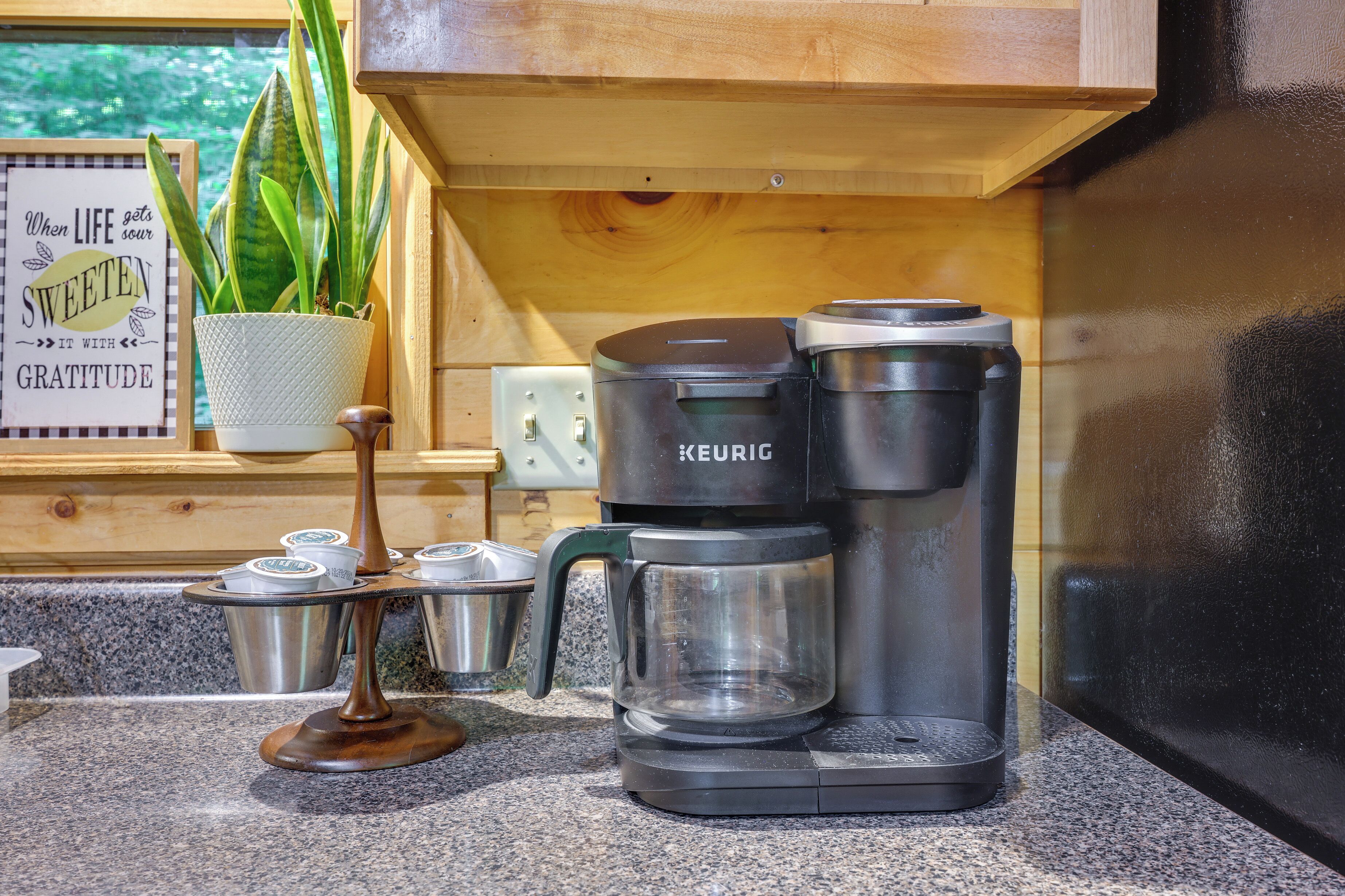 En kaffemaskine på en bakkekløvet bordplade med en potteplante og et opmuntrende skilt i baggrunden.