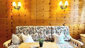A wooden wall with two wall-mounted lamps, a floral-patterned sofa with cushions, and a wooden table with a magazine and a potted plant. - Vacation home Anni - a retreat to relax and enjoy nature at Lake Tegernsee (Kreuth)