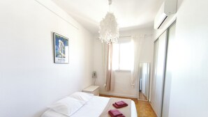 A bedroom with a bed, a bedside table, a hanging light fixture, a window with curtains, and a wall-mounted air conditioner.