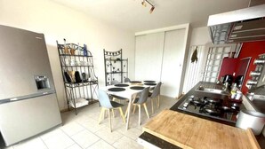 A kitchen with a gas stove, a refrigerator, a dining table with chairs, and a metal shelf unit.