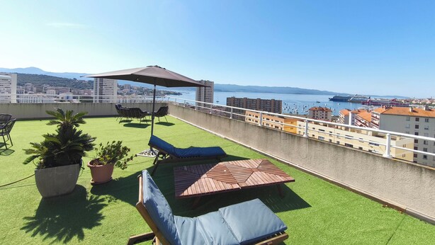 A rooftop terrace with artificial grass, lounge chairs, a wooden table, and a pergola with an umbrella.