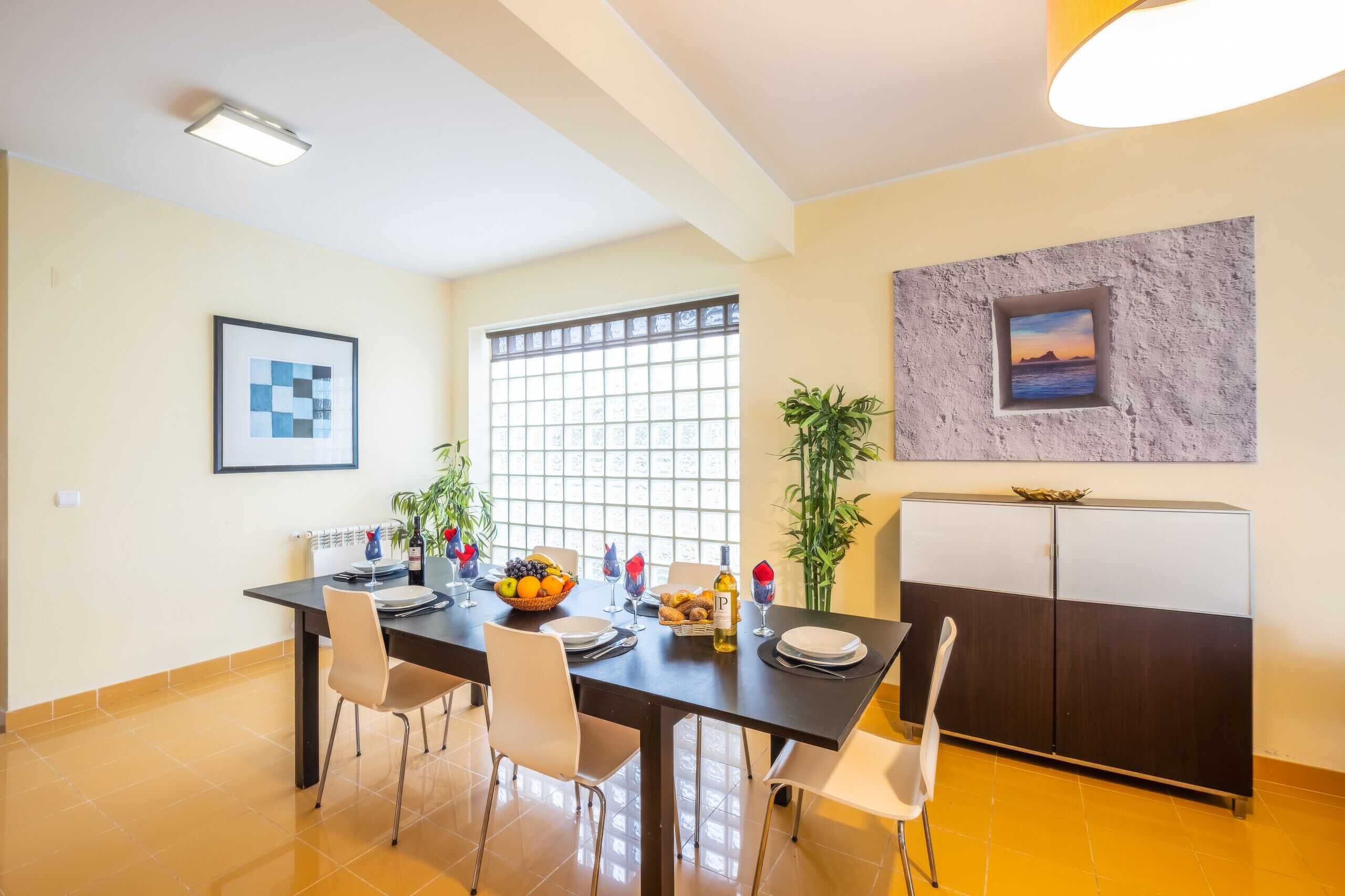 A dining area with a table set for four, chairs, a buffet table with fruits and a bottle of wine, and a refrigerator.