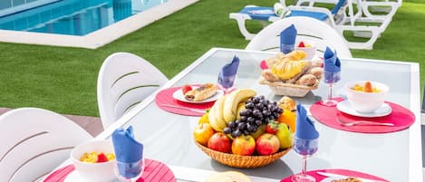 A poolside dining setup with a table set for two, featuring a fruit bowl, bread, and wine glasses.