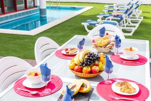 Un coin repas aménagé au bord de la piscine, avec une table dressée pour deux personnes, comprenant un panier de fruits, du pain et des verres à vin.
