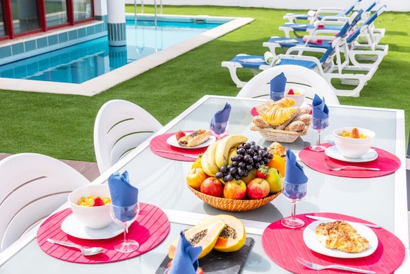 A poolside dining setup with a table set for two, featuring a fruit bowl, bread, and wine glasses.