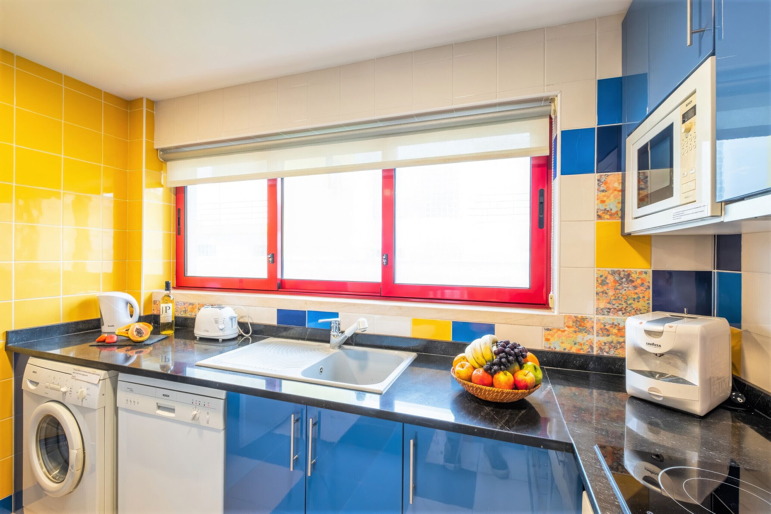 A kitchen with a washing machine, a sink, a microwave, and a fruit bowl on the counter.