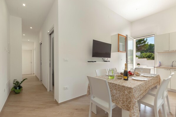 Un espace repas moderne avec une table dressée pour deux personnes, un téléviseur fixé au mur et une vue sur l’extérieur grâce à une grande fenêtre.