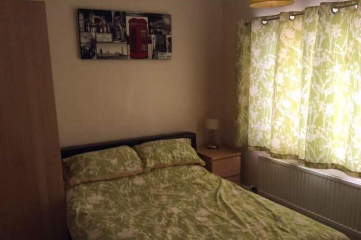 Un dormitorio con cama, mesita de noche, lámpara, ventana con cortinas e imagen colgada en la pared.