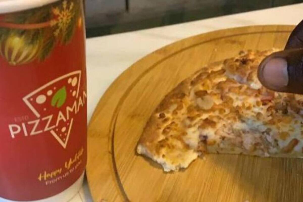 Una porción de pizza sobre una tabla de madera, junto a un vaso rojo con el logotipo de una pizzería.