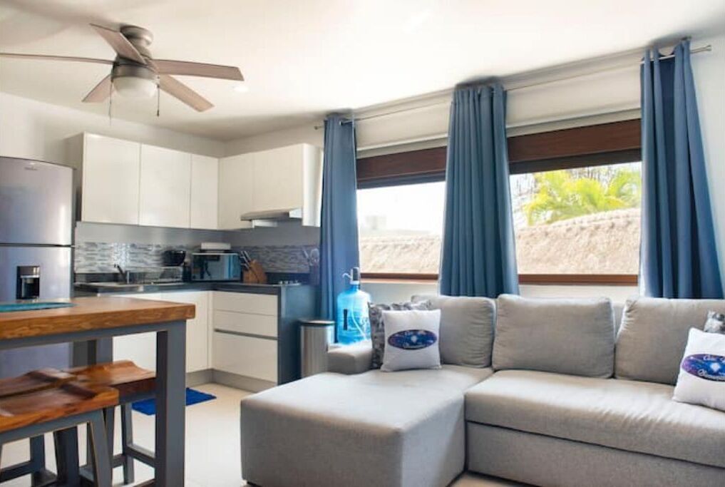 Eine moderne Küche mit weißen Schränken, einem eingebauten Backofen und einer Kaffeemaschine. Ein Essbereich mit einem Holztisch und Bänken lädt zum Verweilen ein. Ein graues Sofa steht in der Nähe eines Fensters mit blauen Vorhängen.