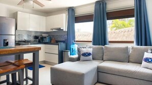 Una cocina moderna con armarios blancos, horno empotrado y cafetera. Hay una zona de comedor con una mesa de madera y bancos. Un sofá gris se encuentra cerca de una ventana con cortinas azules.