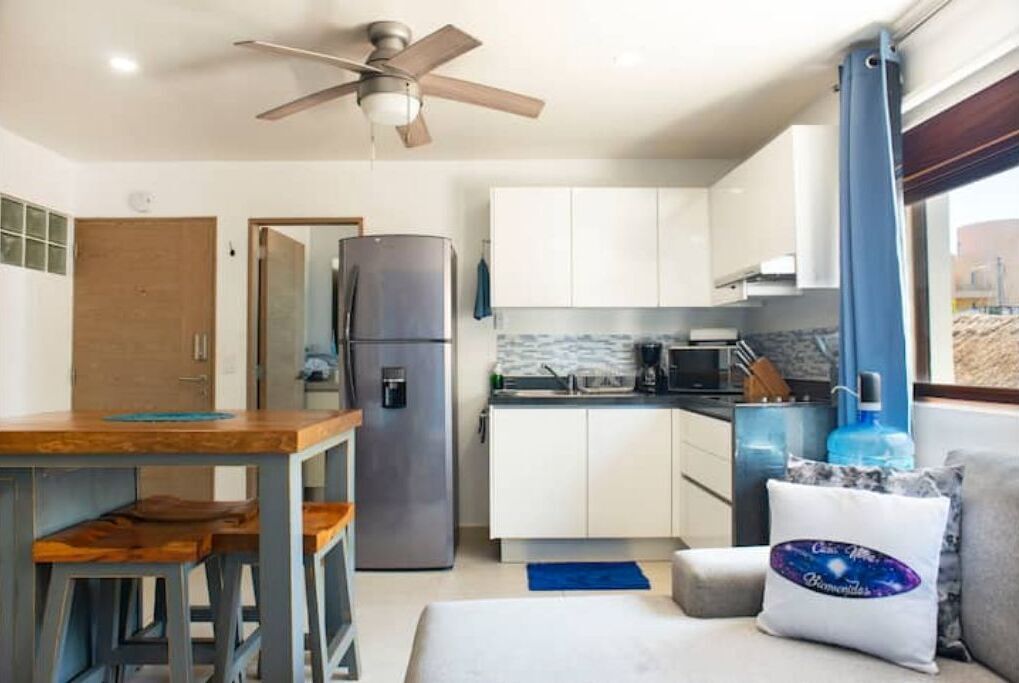 Cocina compacta con refrigerador, microondas e instalación de lavamanos. Hay un ventilador de techo, una mesa de madera con sillas y una ventana con cortinas.