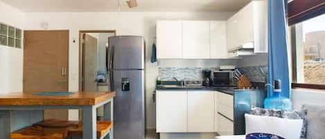 Cocina compacta con refrigerador, microondas e instalación de lavamanos. Hay un ventilador de techo, una mesa de madera con sillas y una ventana con cortinas.