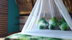 Een bed met een hemelbed en groene kussens, in een kamer met rieten dak en houten balken.