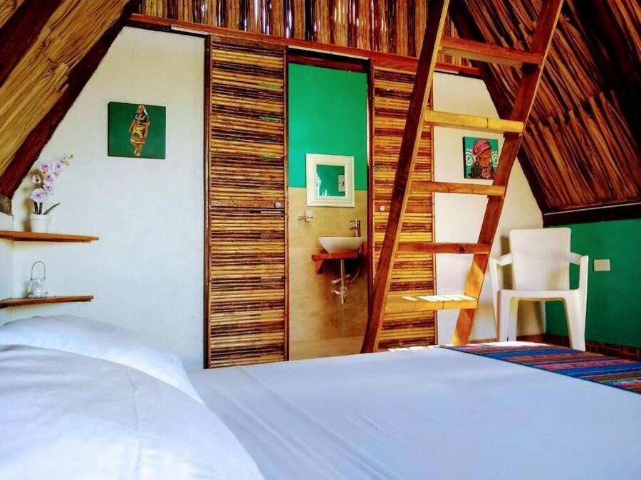 Una camera da letto con soffitto spiovente, un letto, una sedia bianca, un tavolino e una porta verde.