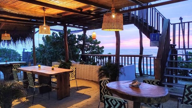 Een terras buiten met houten tafels en stoelen, een trap en uitzicht op de oceaan tijdens de zonsondergang.