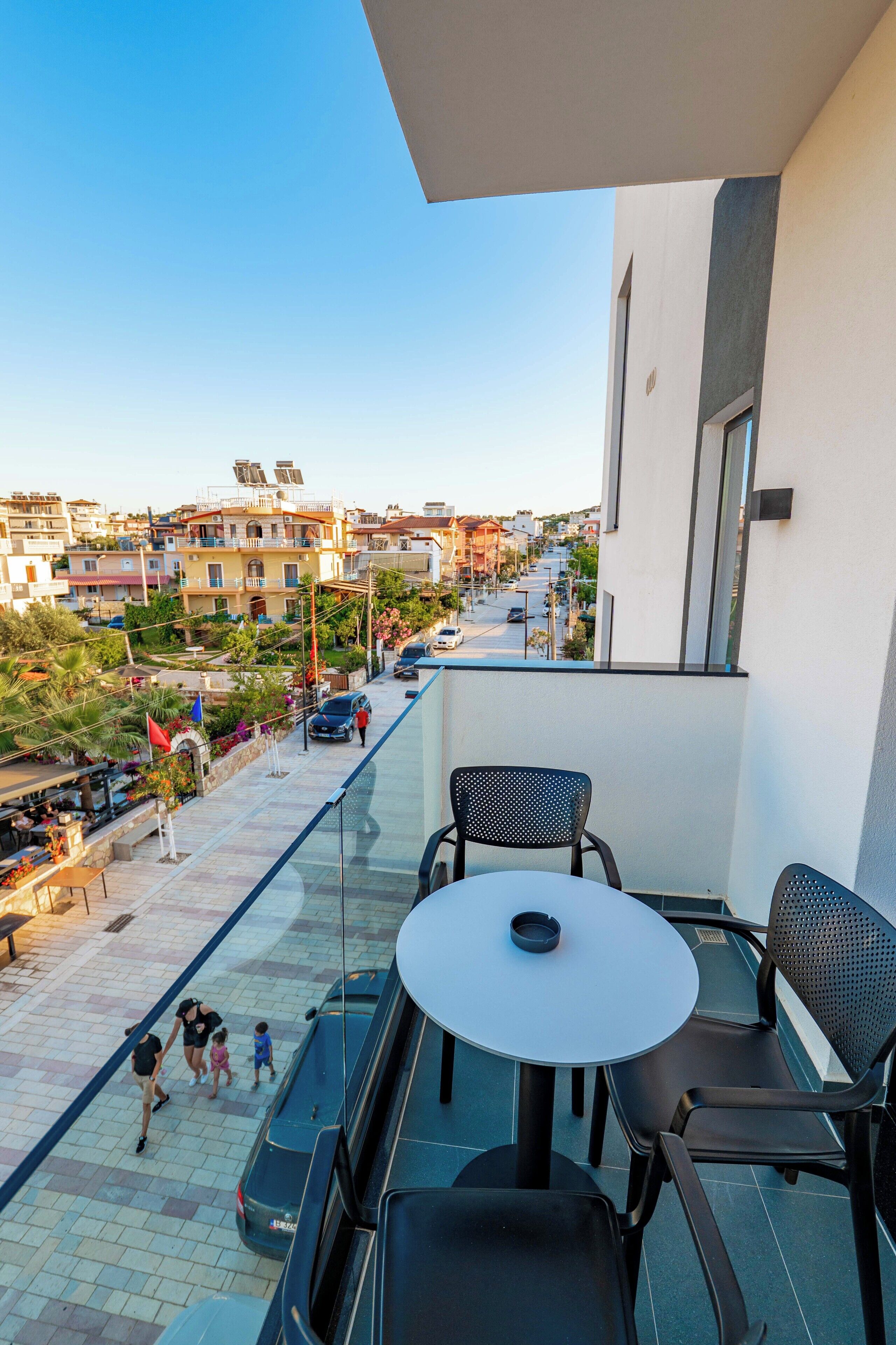 A balcony with a glass railing overlooking a street with cars and buildings. There is a table and two chairs on the balcony.